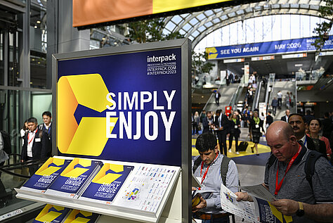 A trade fair booth of Interpack Alliance with a large blue and yellow sign saying 'SIMPLY ENJOY'. Flyers with the logo are displayed on a stand, and visitors are visible in the background at the event.