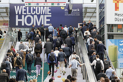 A large crowd of people ascending escalators toward a sign that reads 'WELCOME HOME' at the Interpack event entrance.