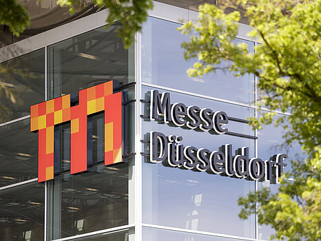 The exterior of a modern glass building with the logo 'Messe Düsseldorf' prominently displayed on the facade, partially framed by green tree branches.