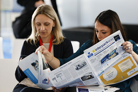 Two women seated and reading an issue of the 'interpack news' newspaper, focusing on event-related content.