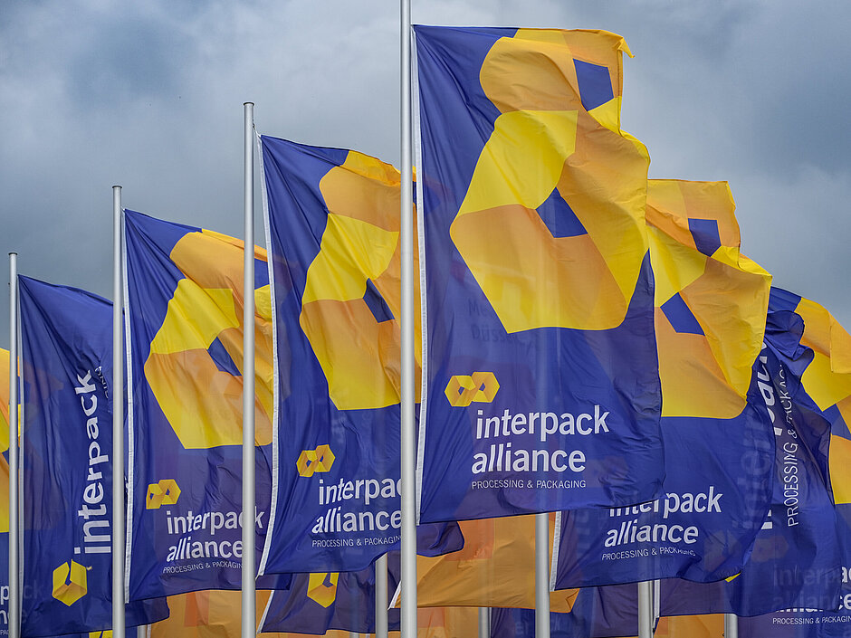 Several blue flags waving, featuring the yellow-orange logo and the text 'interpack alliance – Processing &amp; Packaging' against a cloudy sky.